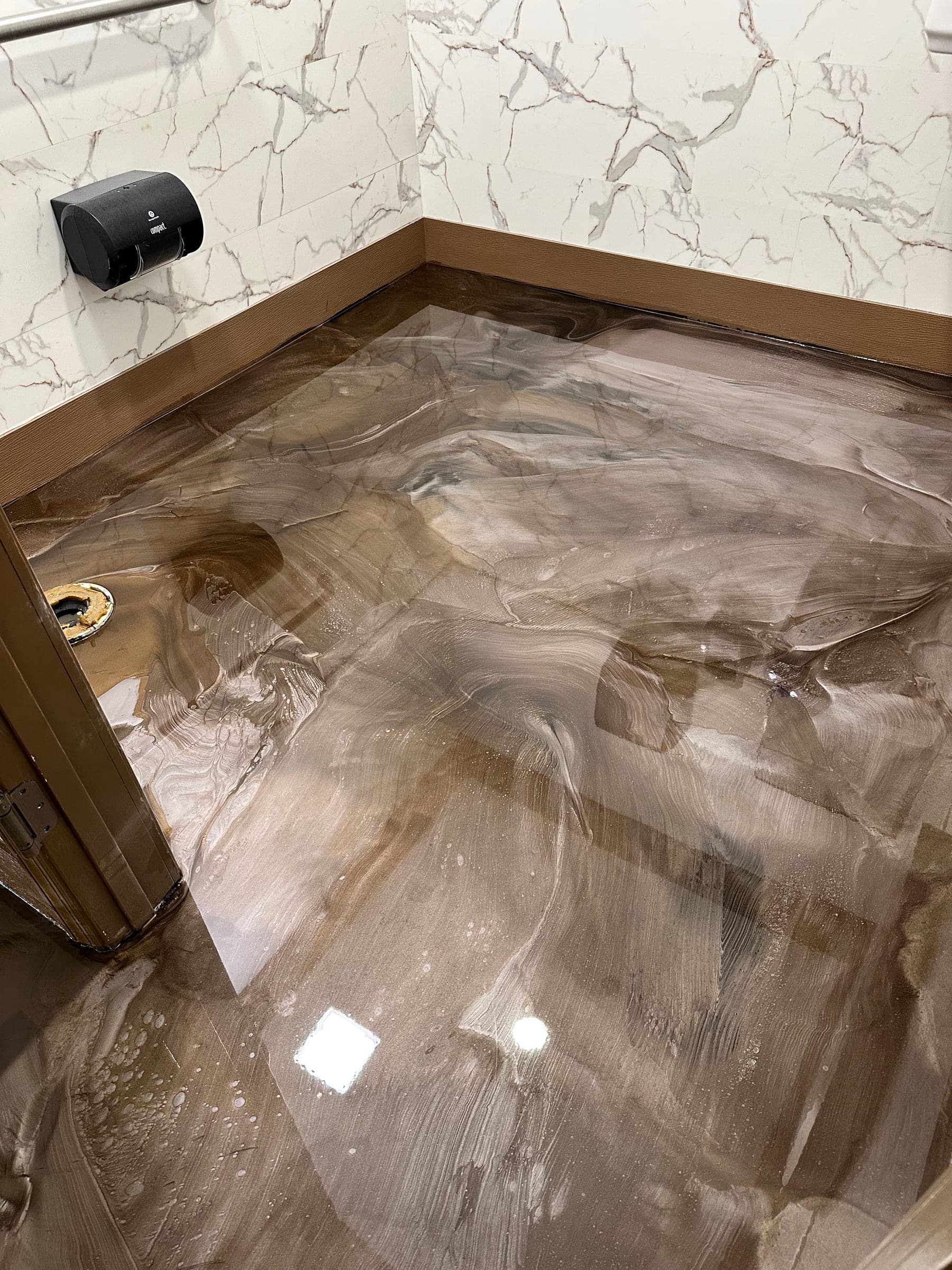 Glossy brown and grey marbled metallic epoxy floor in a commercial restroom with white marble-look tile walls.