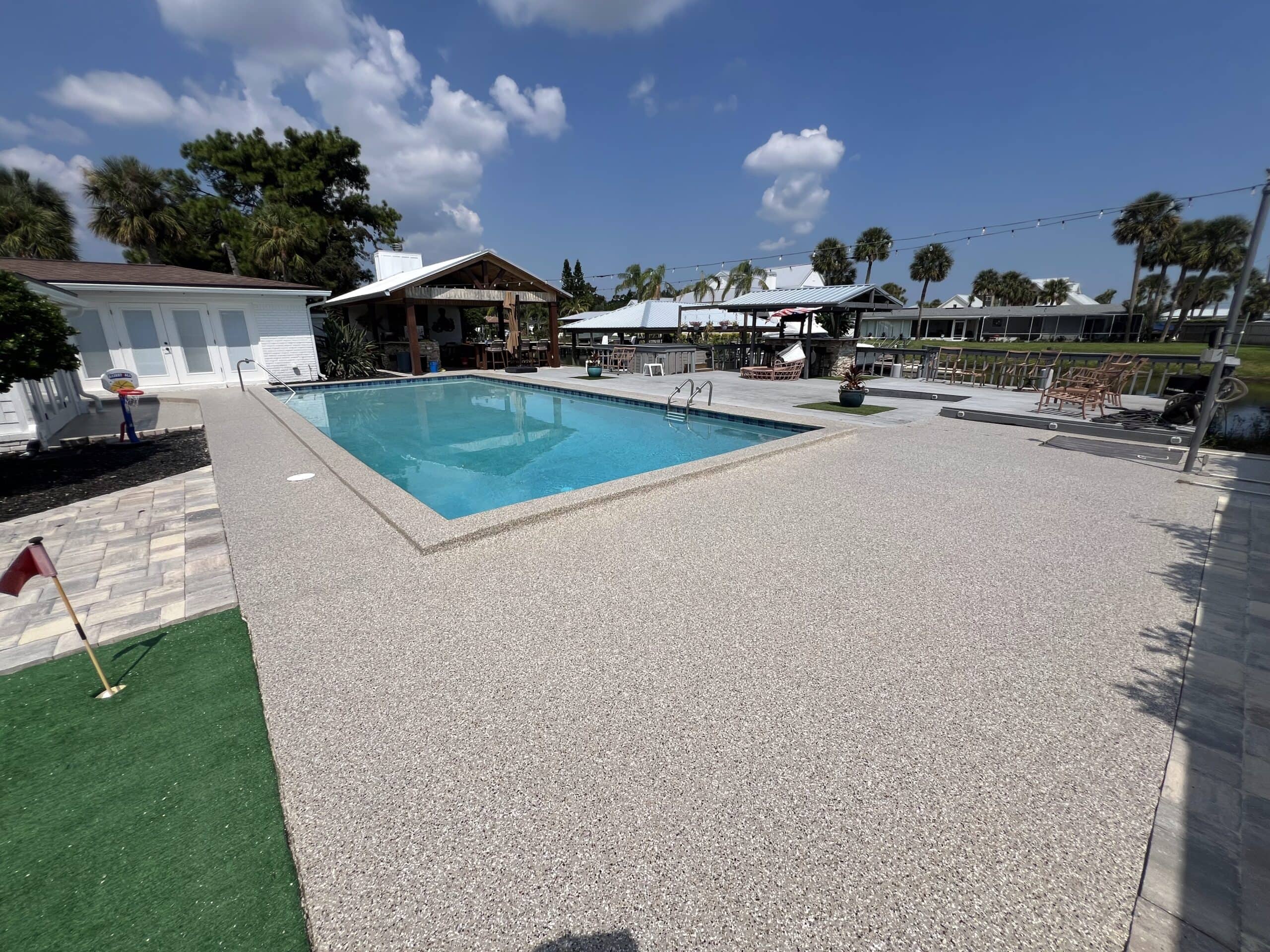 Light grey speckled pool deck surrounding a rectangular outdoor swimming pool with seating and palm trees nearby.